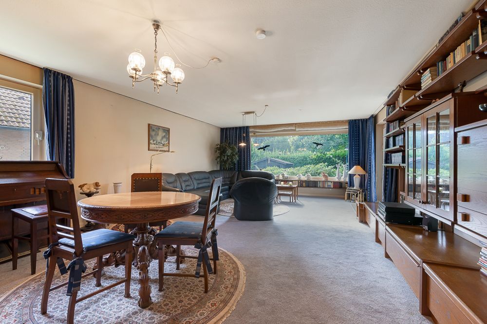 Wohnzimmer mit rundem Holztisch, Stühlen, Ledersofa und großer Fensterfront mit Blick ins Grüne.