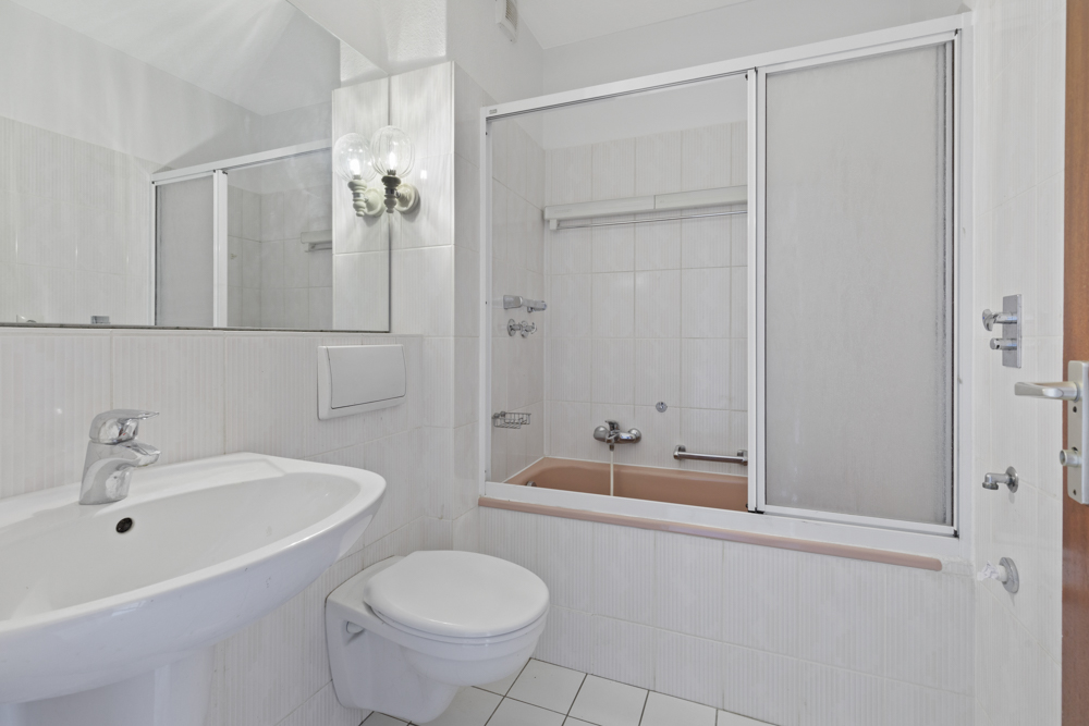 Helles Bad mit Waschbecken, Wand-WC und Badewanne mit Glas-Duschabtrennung.