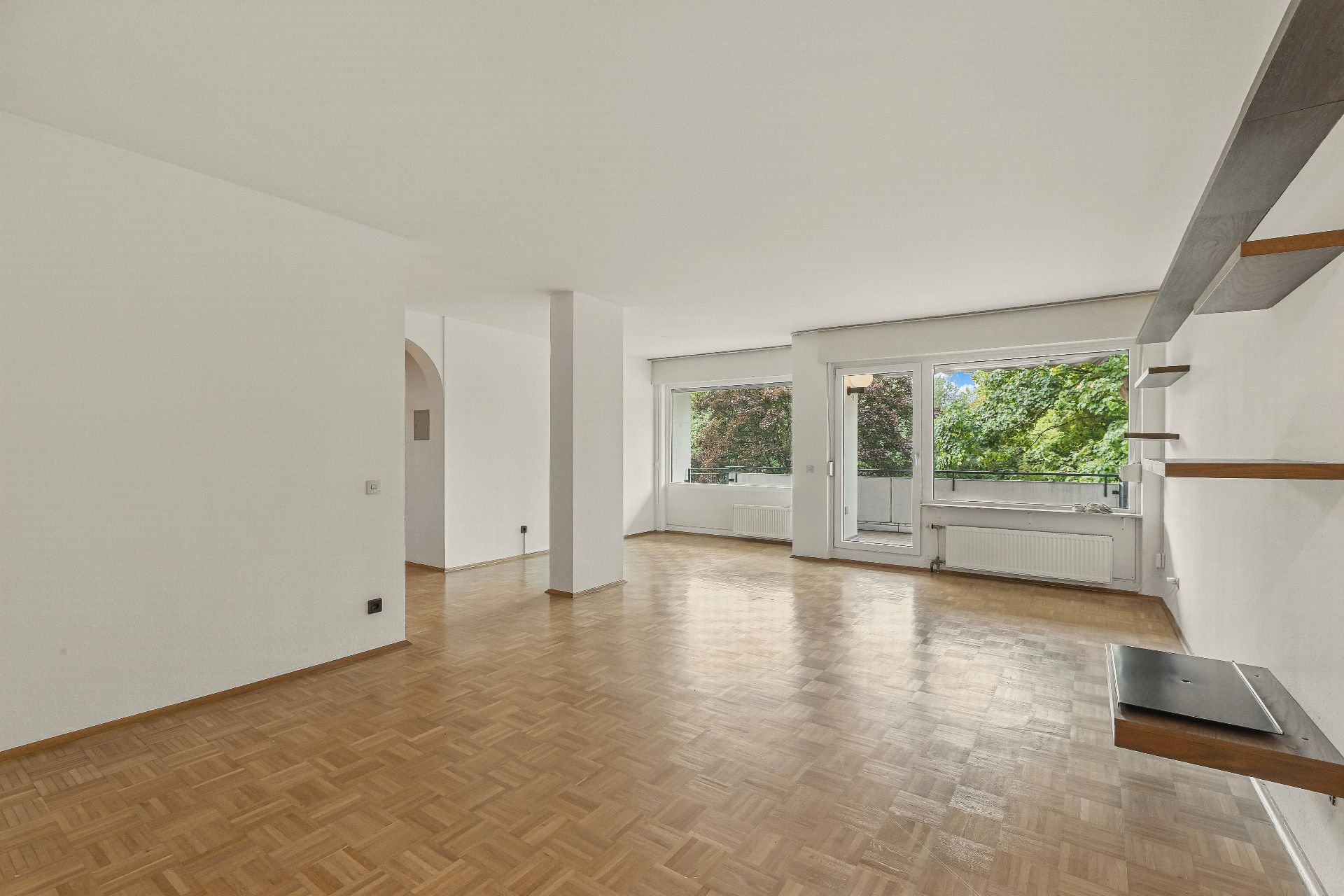 Helles Wohnzimmer mit Parkettboden, großen Fenstern und Regalen an der Wand.