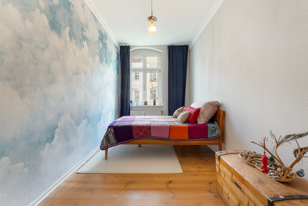 Schmales Schlafzimmer mit Holzbett, bunter Decke, Wolkentapete und Holzkiste auf Holzboden.