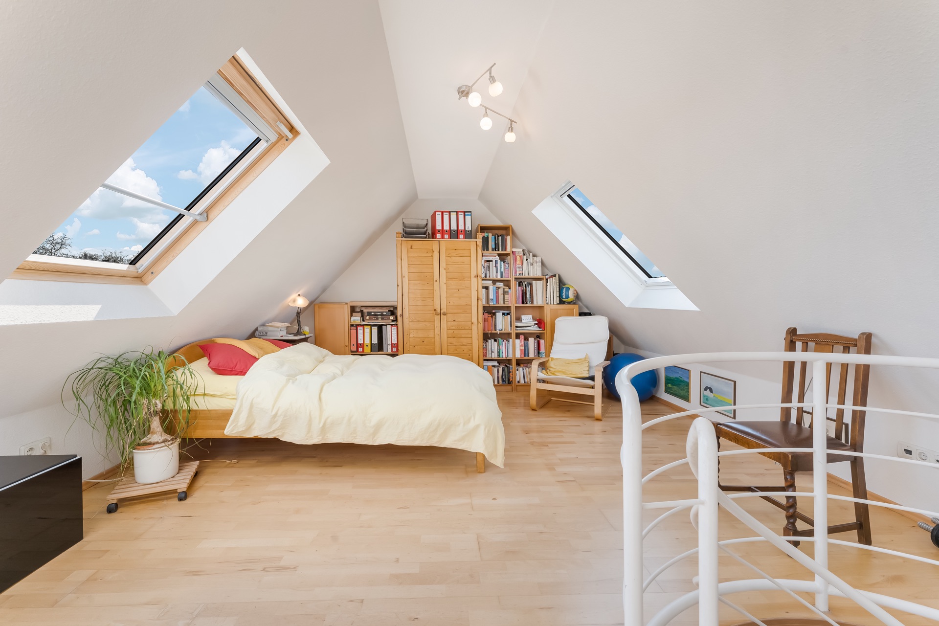 Helles Dachzimmer mit Bett, Sessel, Bücherregal, Pflanzen und zwei Dachfenstern.