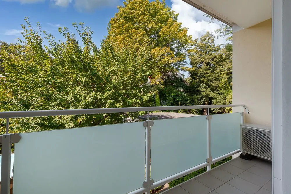 Balkon mit Glasgeländer und Blick auf grüne Bäume.
