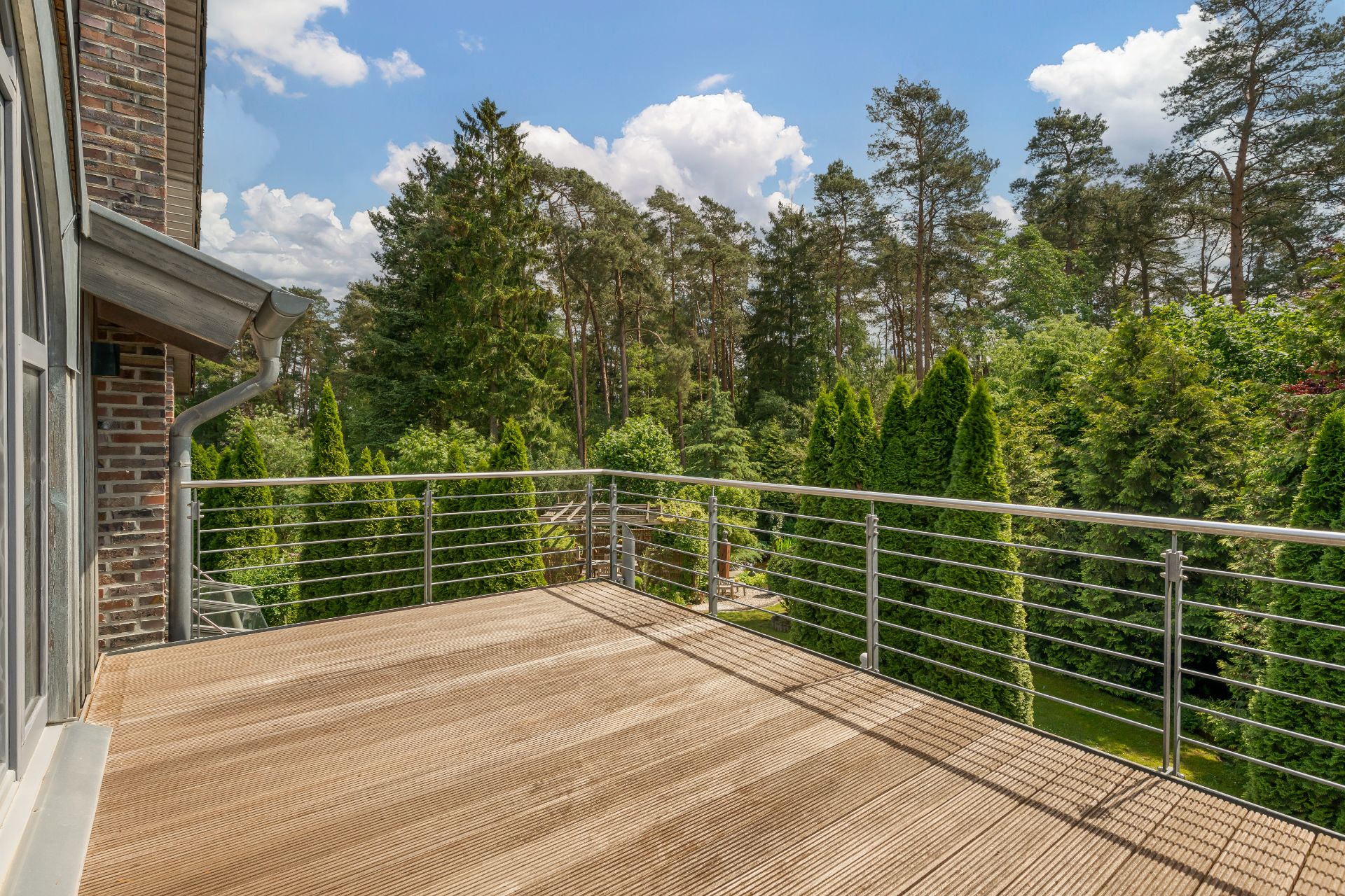 Großer Balkon mit Holzboden und Metallgeländer, Blick auf grüne Bäume und blauen Himmel.