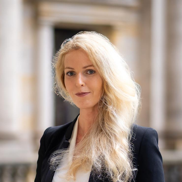 Eine Frau mit langen blonden Haaren steht in einem Innenhof und trägt einen schwarzen Blazer.