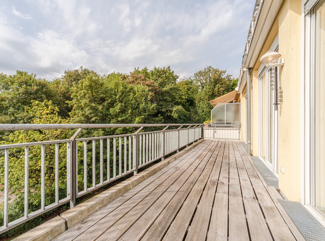 Großer Holzbalkon mit Metallgeländer, Blick auf grüne Bäume, angrenzend an ein gelbes Wohngebäude.