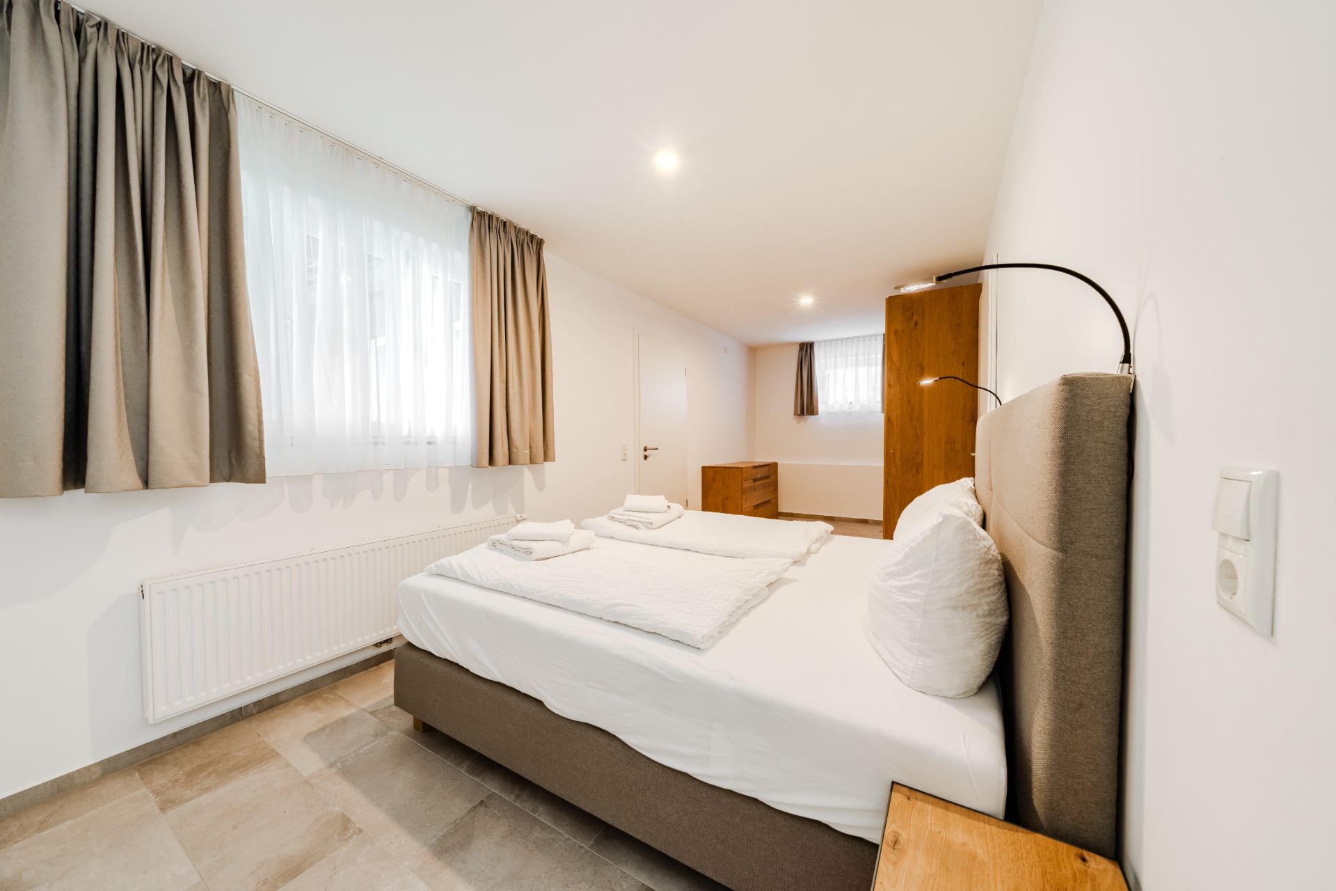 Helles Schlafzimmer mit Doppelbett, zwei Fenstern, Vorhängen und Holzmöbeln.