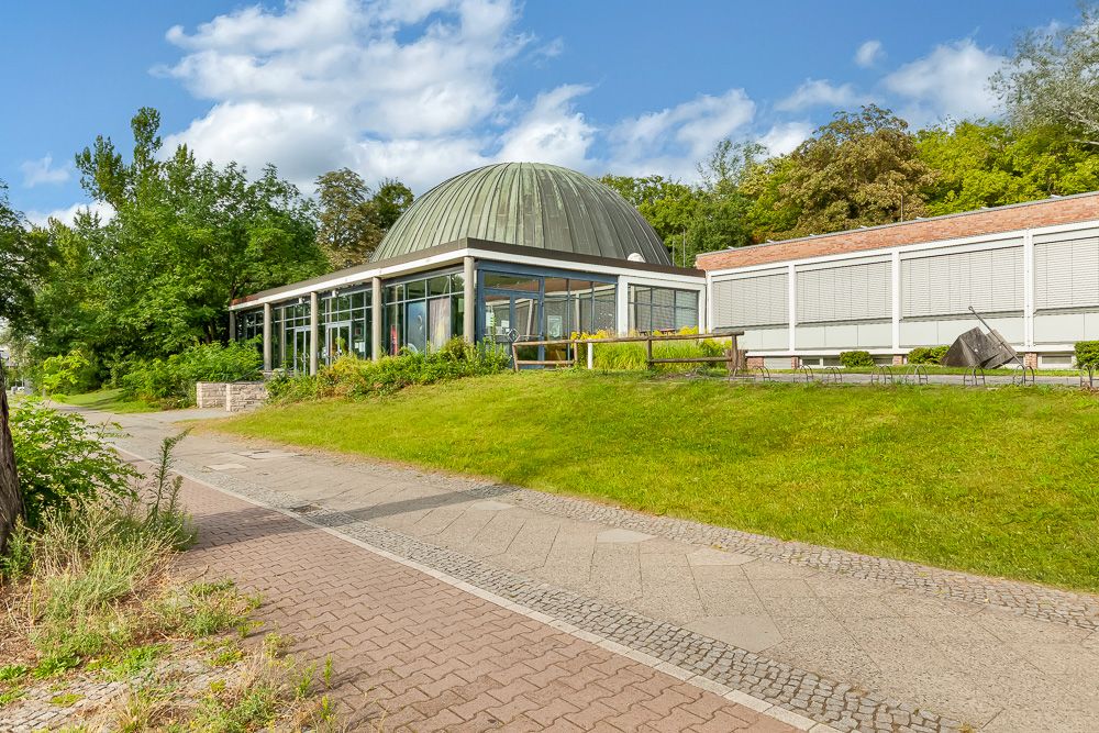 Modernes Gebäude mit Glaskuppel und großen Fenstern, umgeben von viel Grün und Gehwegen.