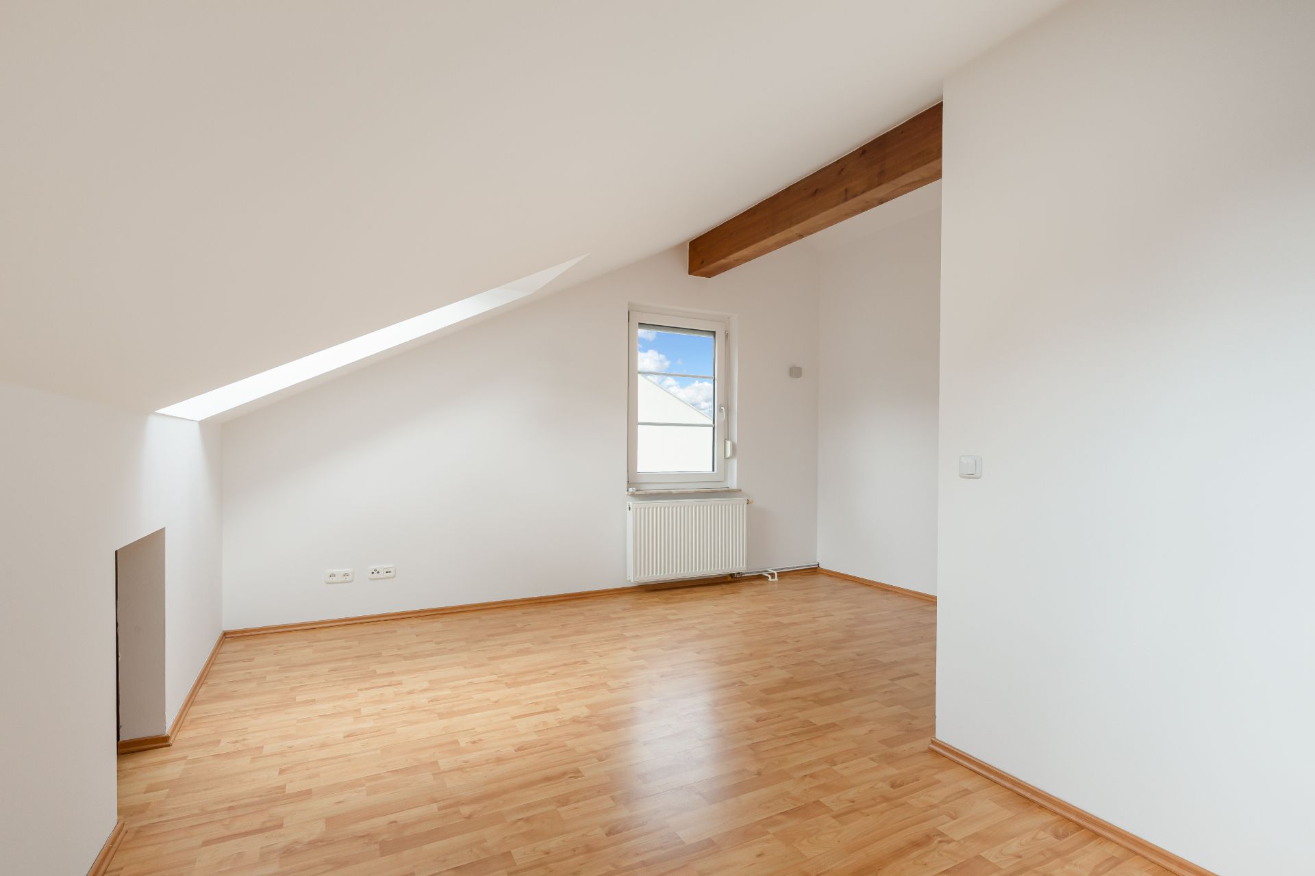 Helles Zimmer mit schräger Decke, Dachfenster, Holzboden und weiß gestrichenen Wänden.