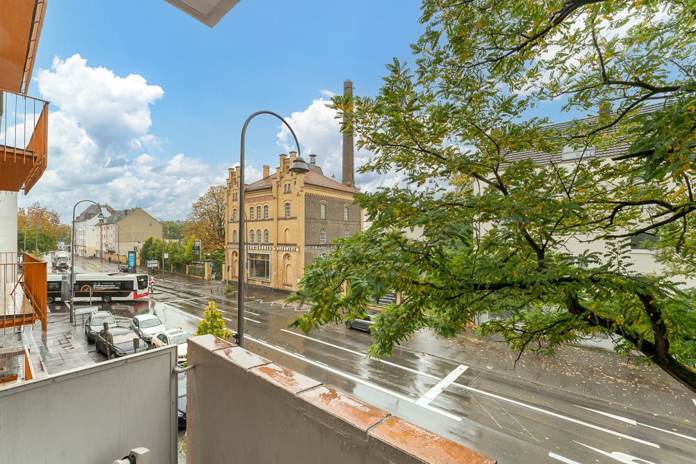 Blick von einem Balkon auf eine nasse Straße mit Autos, Bus, Bäumen und einem Backsteingebäude.