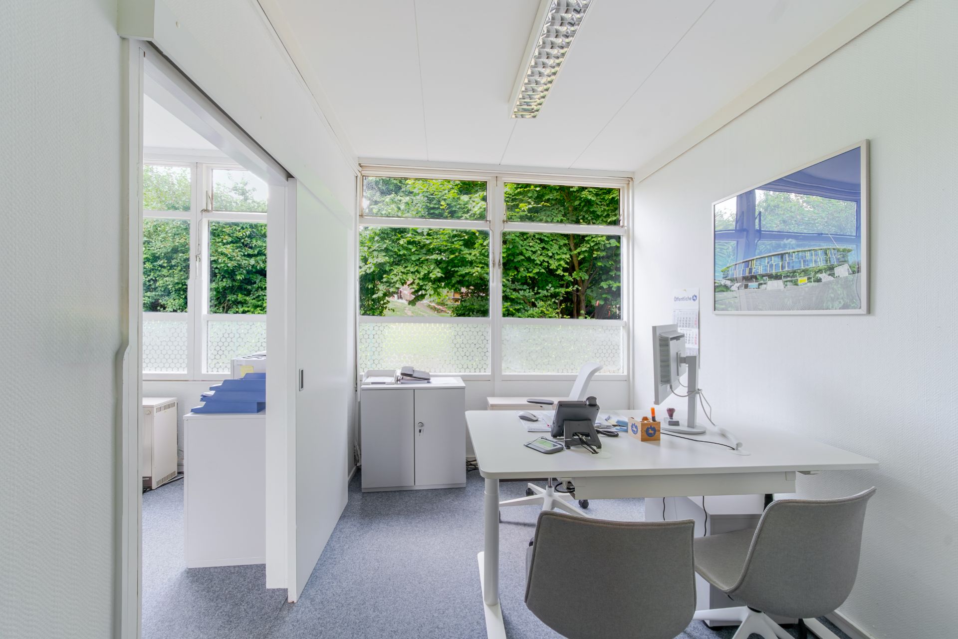 Helles Zimmer mit Schreibtisch, Bürostühlen und großen Fenstern mit Blick ins Grüne.
