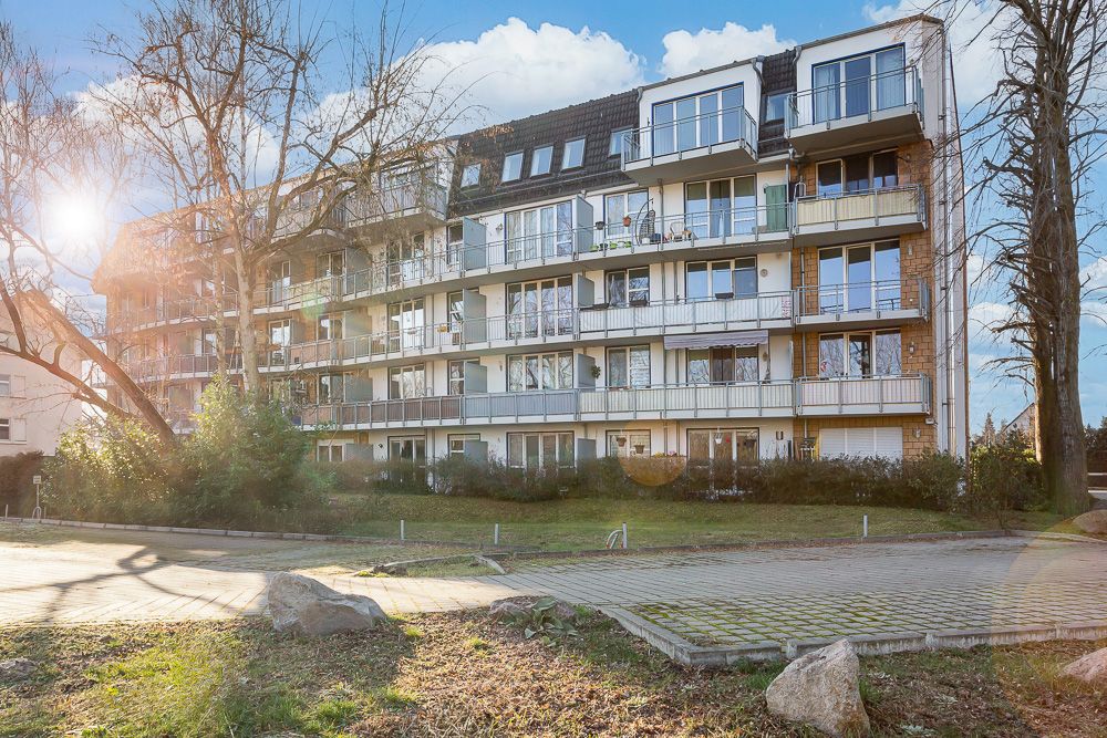 Mehrfamilienhaus mit mehreren Balkonen, Bäumen und gepflastertem Vorplatz bei Sonnenschein.