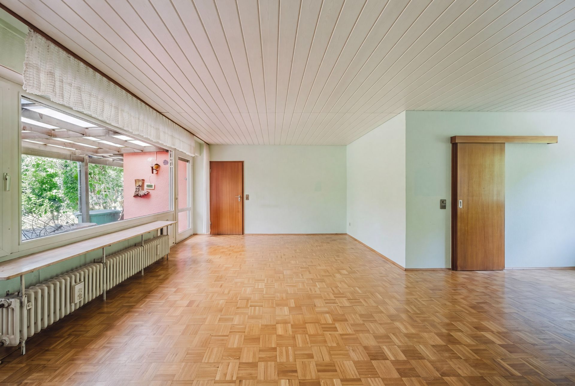 Großes, leeres Zimmer mit Parkettboden, weißen Wänden und großen Fenstern.