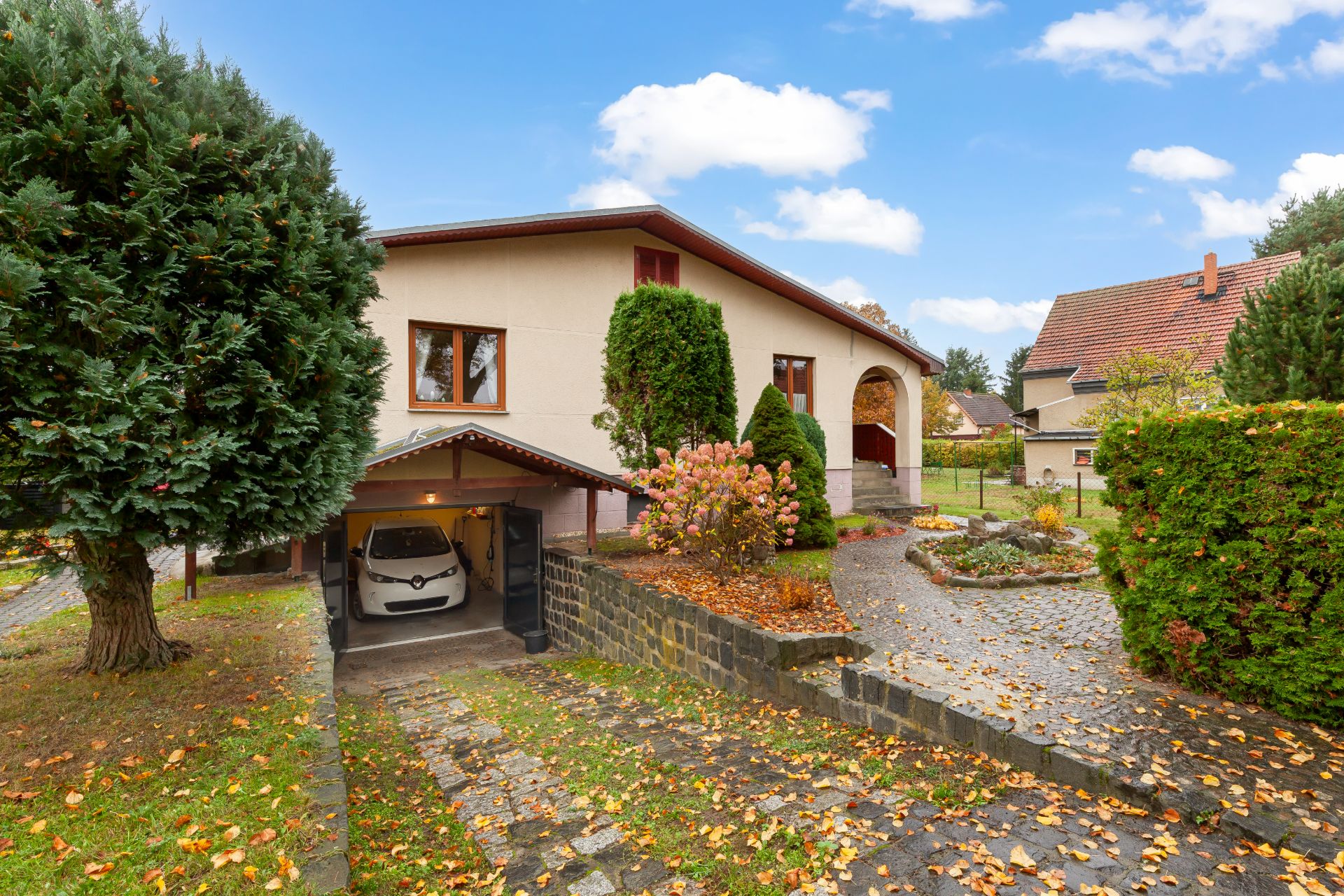 Einfamilienhaus mit Garage, Auto, Garten und Gehweg, umgeben von Bäumen und Herbstlaub.