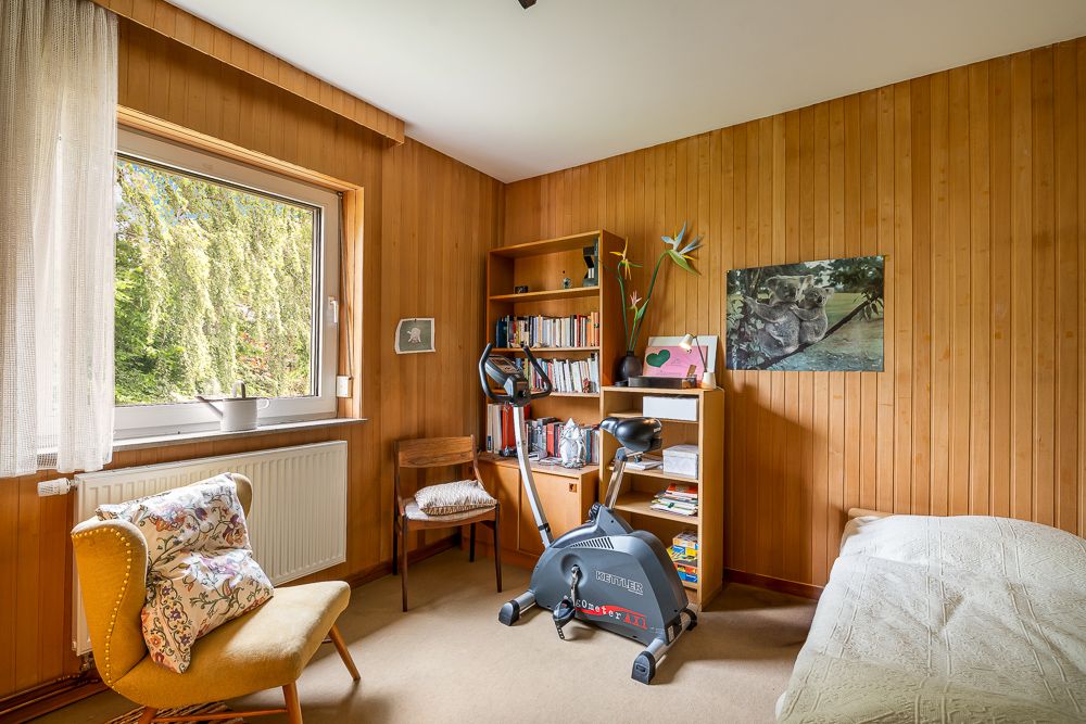 Zimmer mit Holzvertäfelung, Bett, Bücherregal, Stuhl, Sessel, Fenster und Heimtrainer.