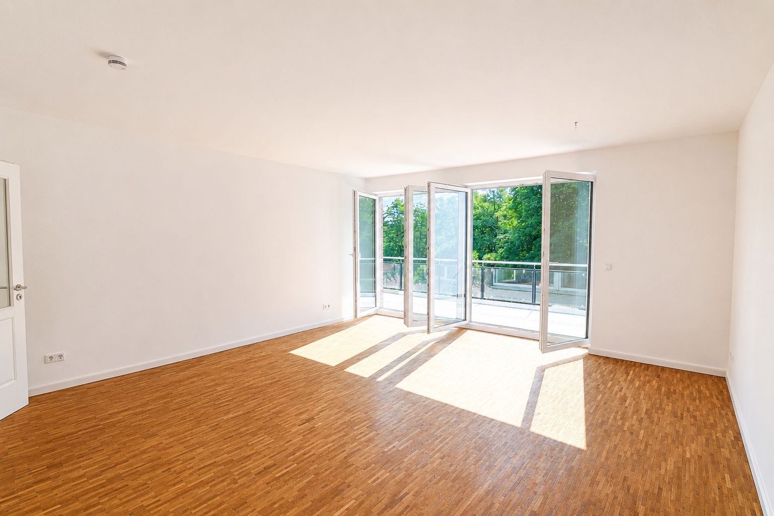 Helles, leeres Zimmer mit Parkettboden und großen Fenstern zu einem Balkon mit Blick auf Bäume.