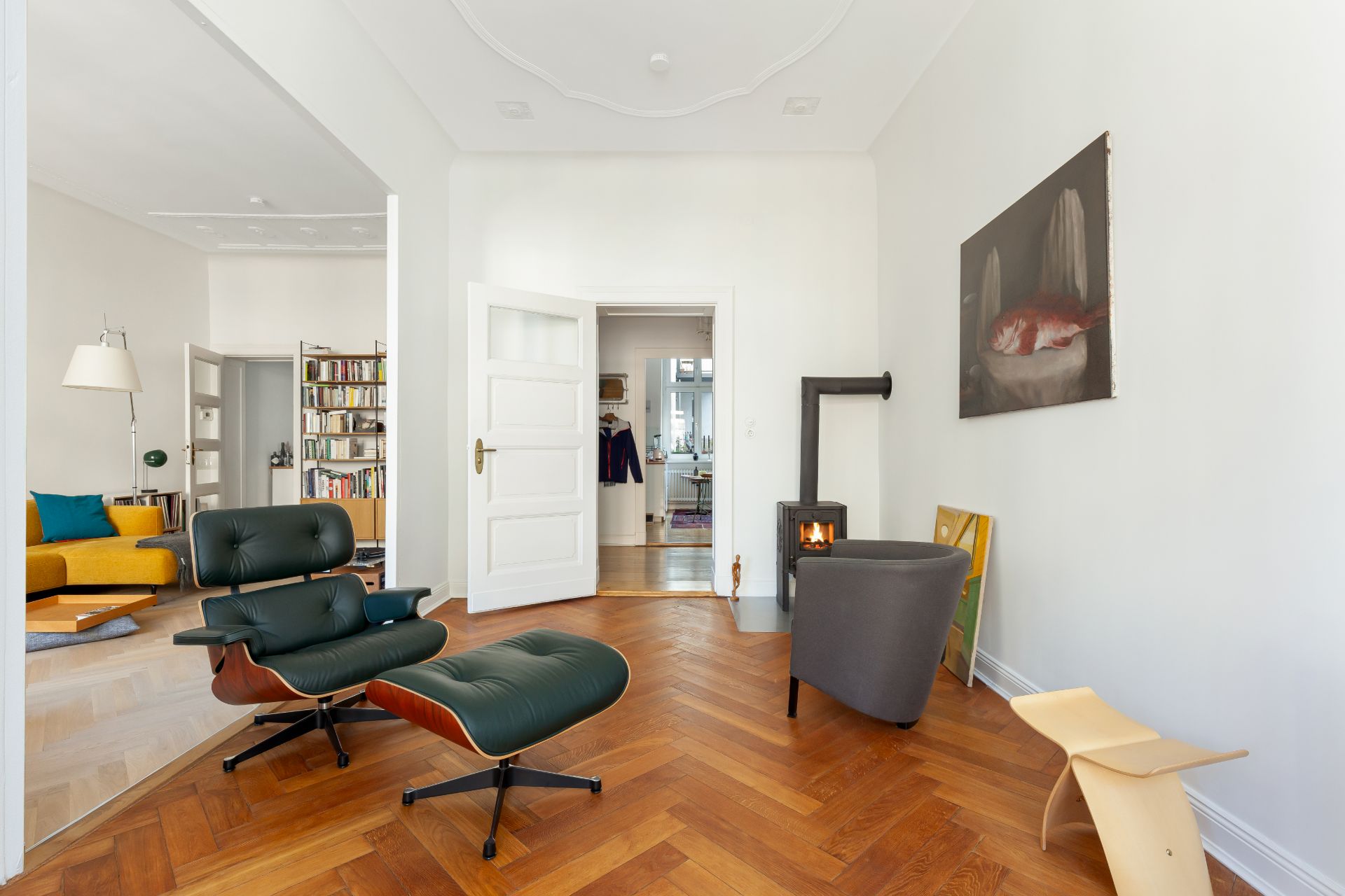 Modernes Wohnzimmer mit Sessel, Ofen, Holzfußboden und Blick in ein zweites Zimmer mit Bücherregal.
