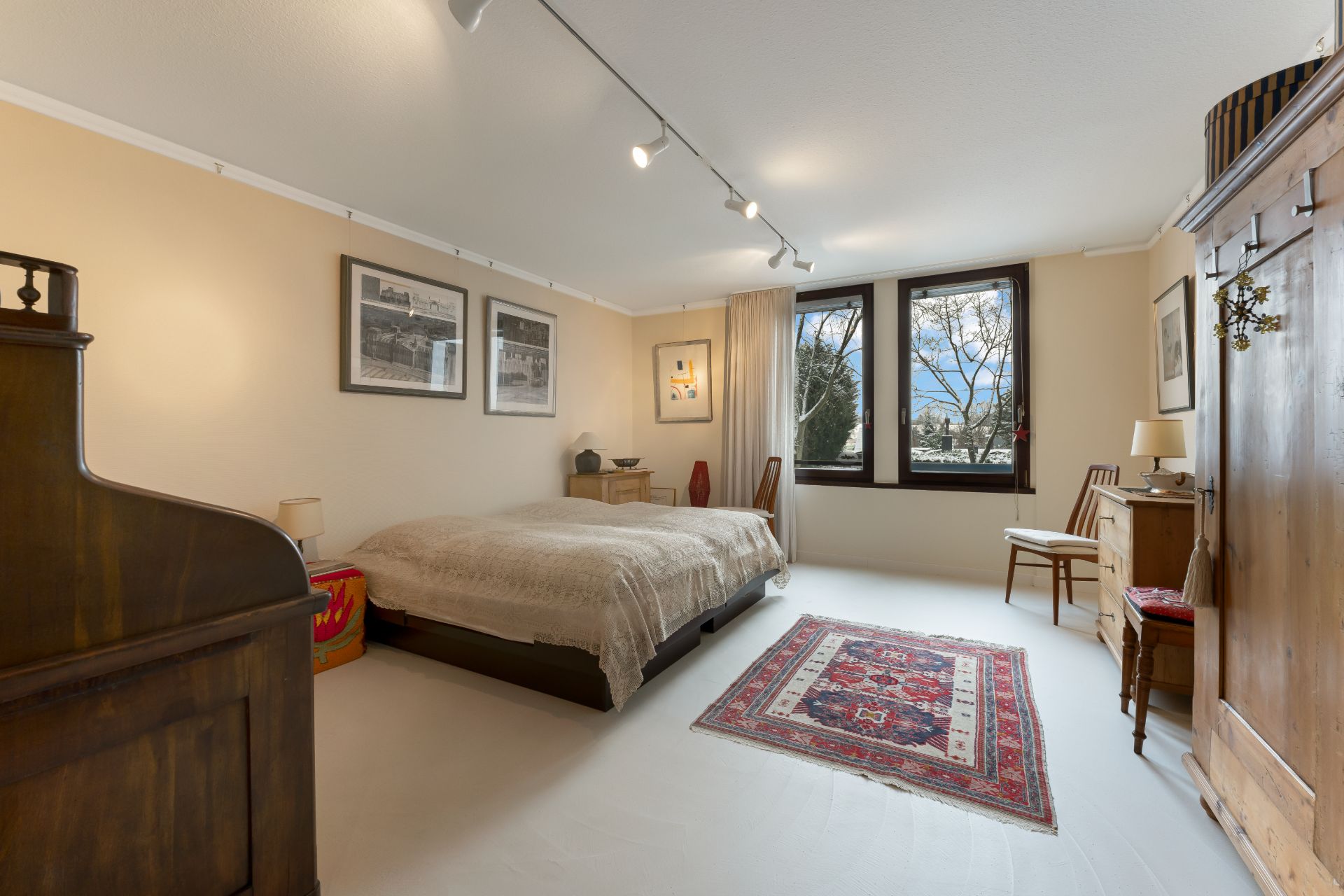 Helles Schlafzimmer mit Doppelbett, Teppich, Holzmöbeln und zwei Fenstern mit Blick nach draußen.