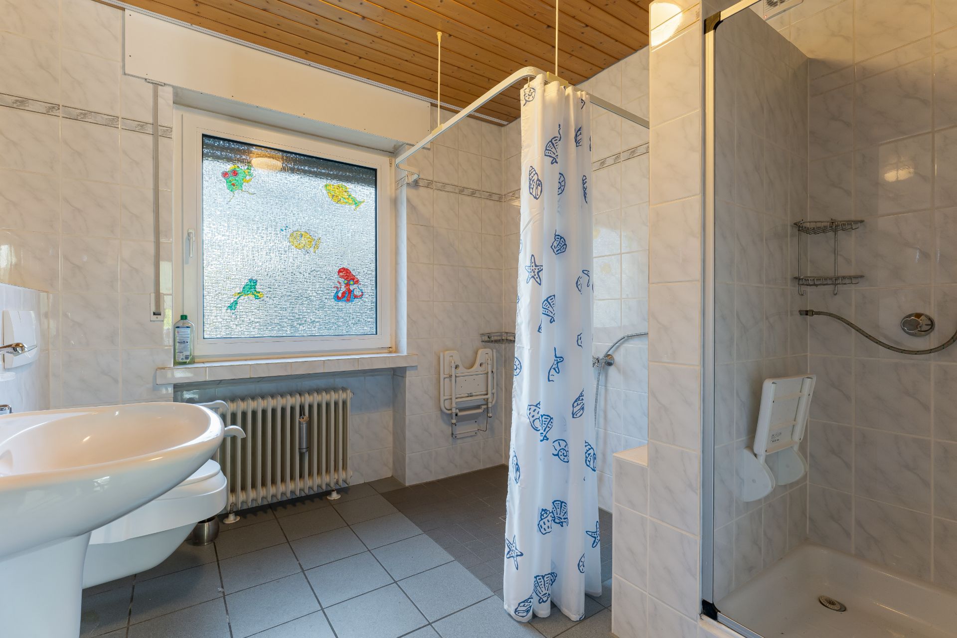 Barrierefreies Badezimmer mit Dusche, Duschvorhang, WC, Waschbecken und Fenster mit bunten Motiven.