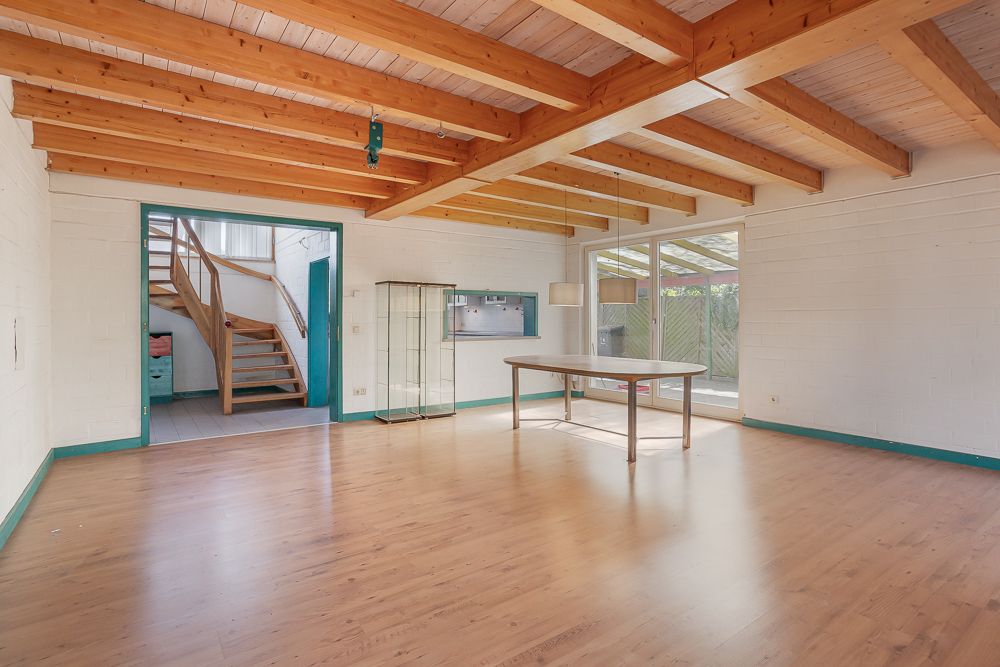 Heller Raum mit Holzbalkendecke, großem Fenster, einem Tisch und einer Treppe im Hintergrund.
