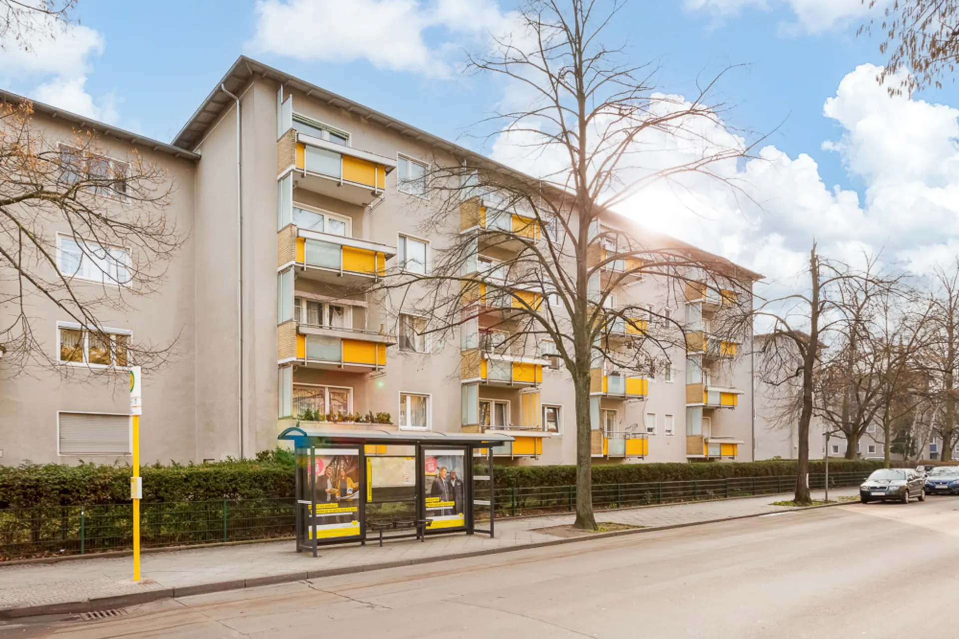 Mehrstöckiges Wohnhaus mit gelben Balkonen und Bushaltestelle davor.