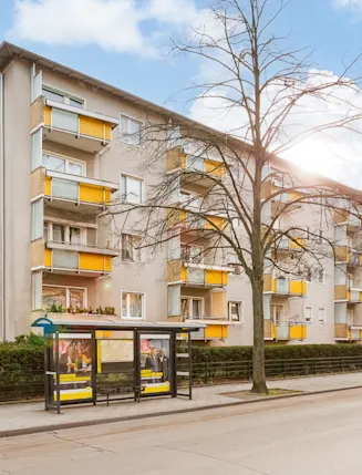 Mehrstöckiges Wohnhaus mit gelben Balkonen und Bushaltestelle davor.