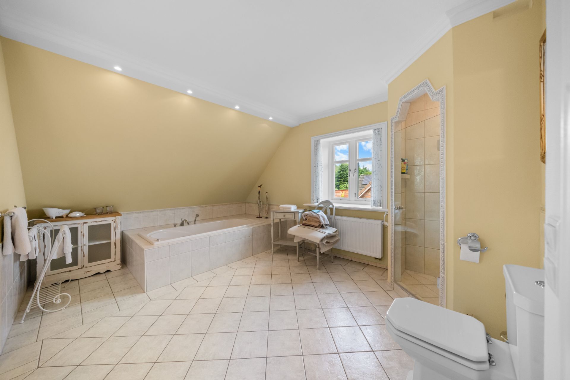 Helles Badezimmer mit Badewanne, Dusche, WC, Fenster und Fliesenboden.