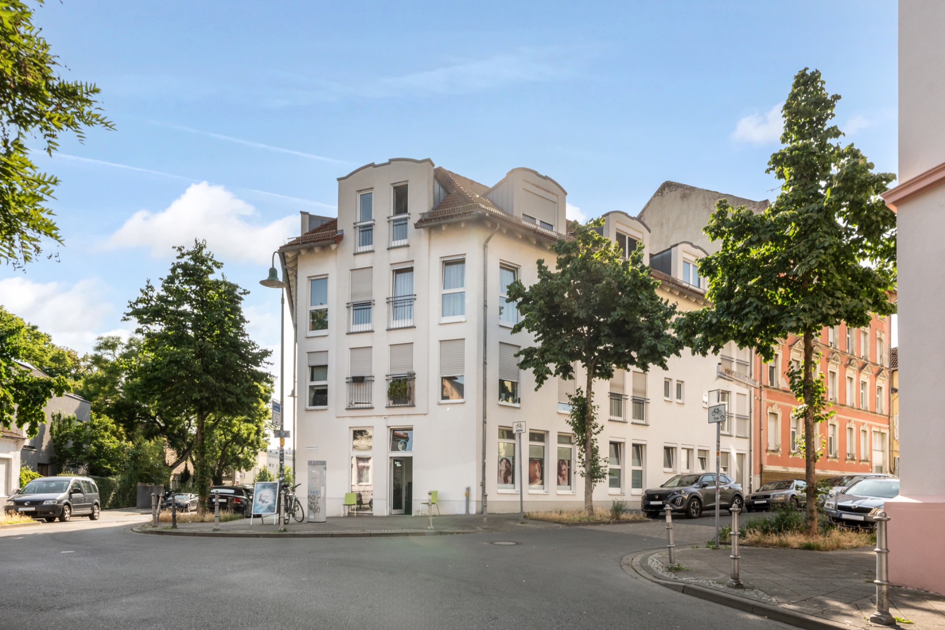 Weiße, mehrstöckige Wohnhaus an einer Straßenecke mit Bäumen und geparkten Autos.