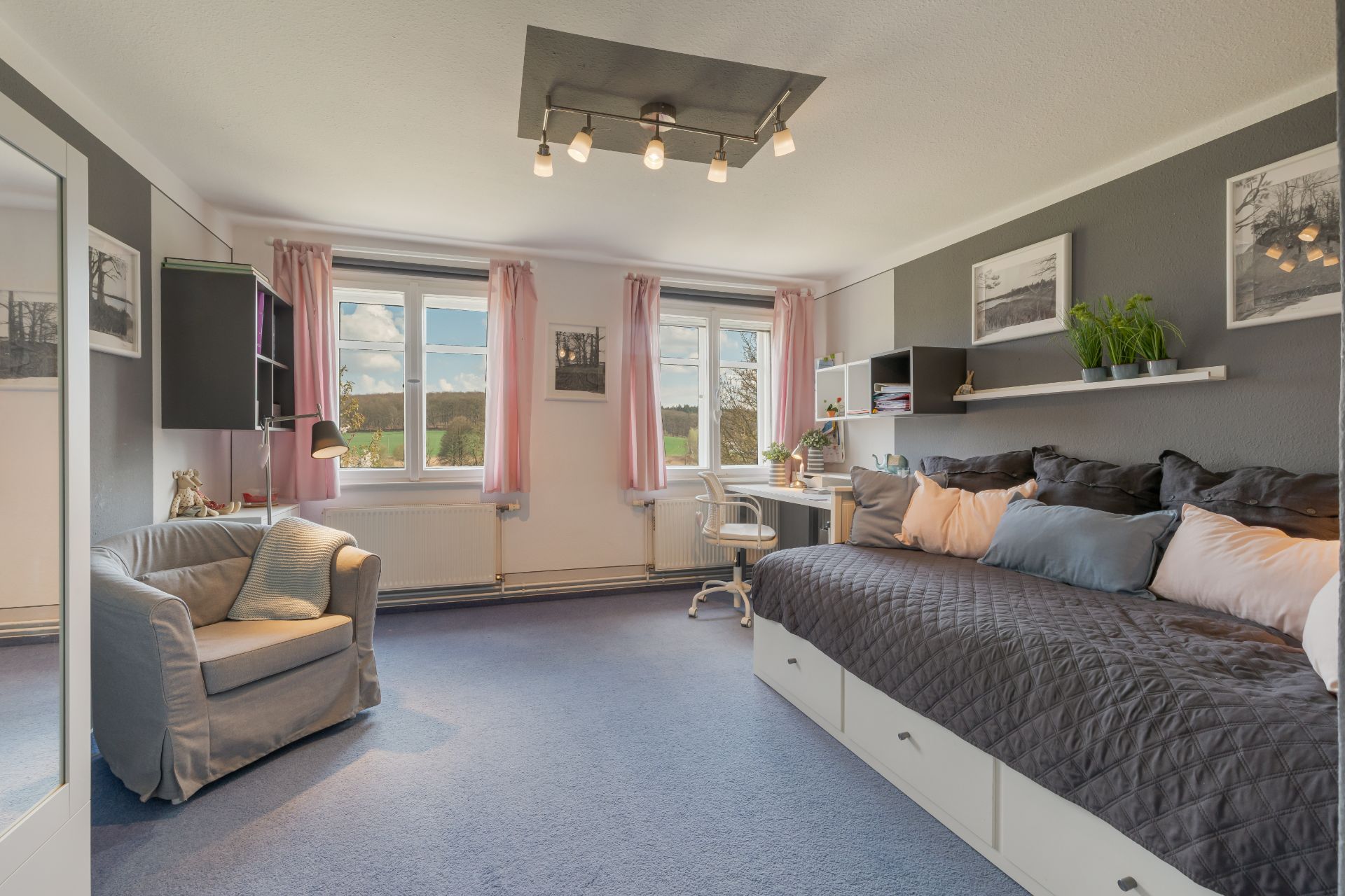 Helles Schlafzimmer mit großem Bett, Sessel, Schreibtisch und Fenstern mit rosa Vorhängen.