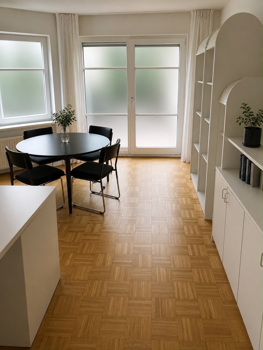 Heller Raum mit rundem Tisch, vier Stühlen, Regal und großen Fenstern mit Milchglas.