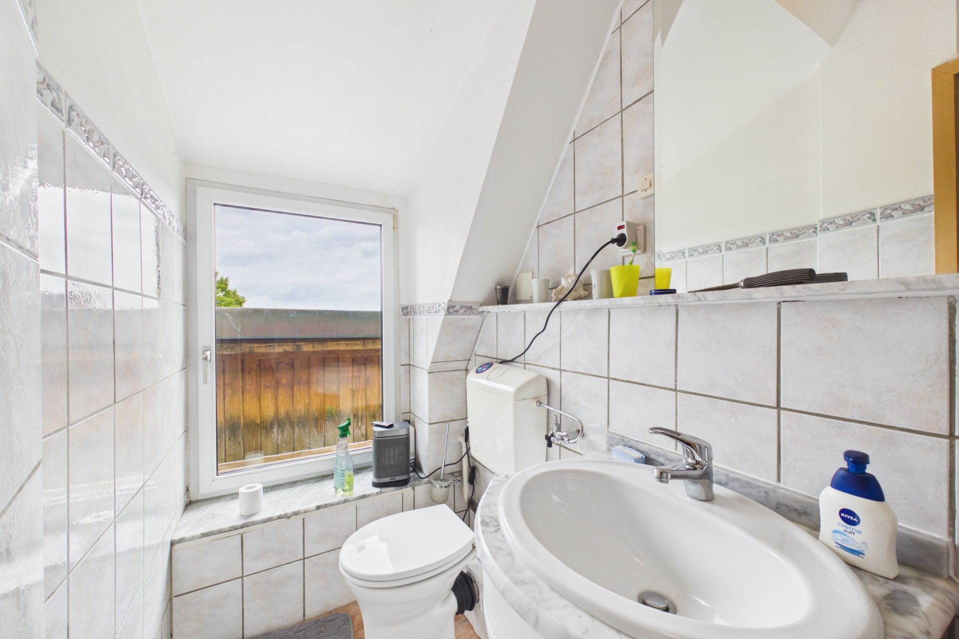 Gefliestes Bad mit Waschbecken, Toilette, Spiegel und Fenster mit Blick nach draußen.