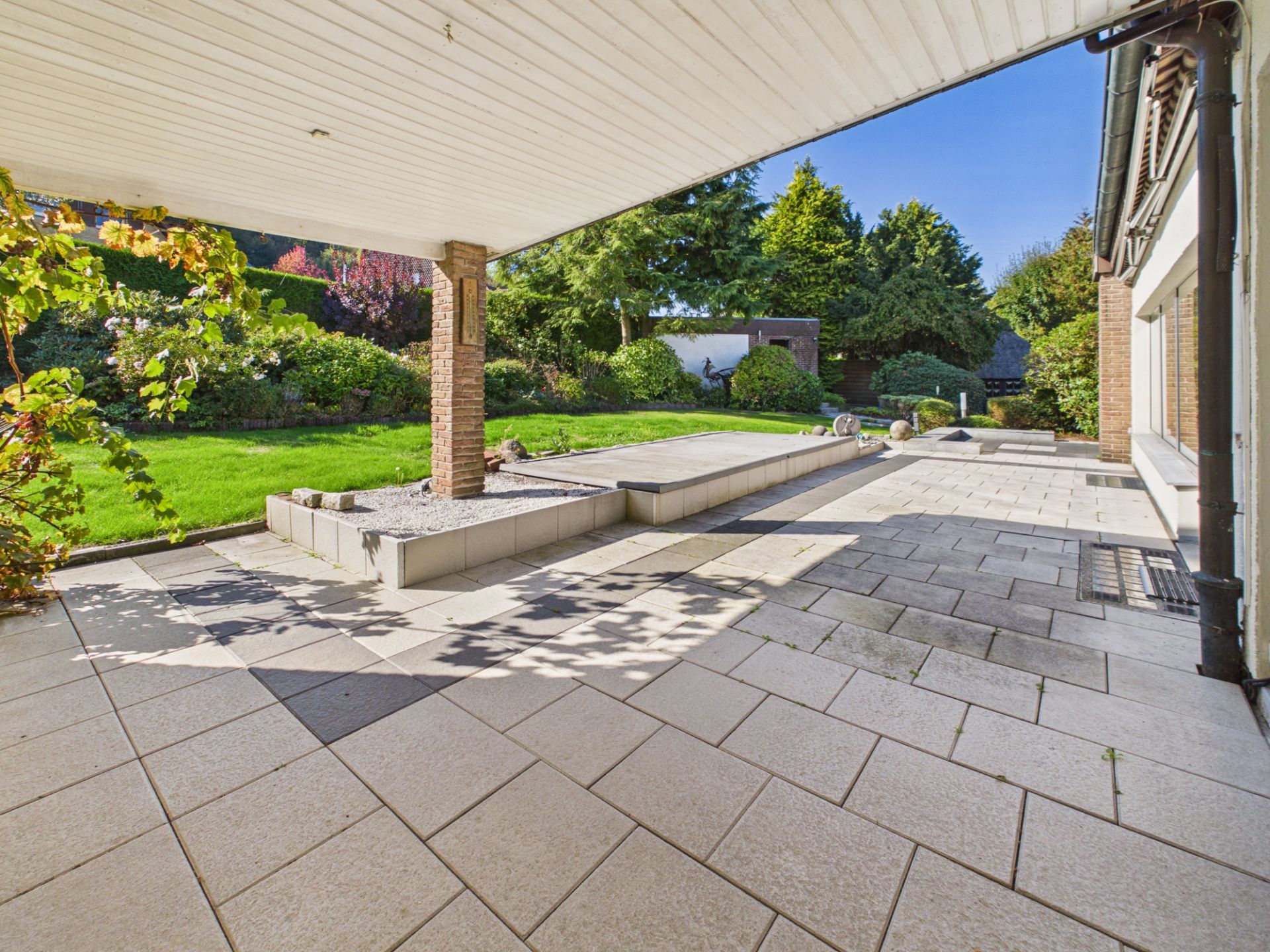 Überdachte Terrasse mit Fliesenboden, angrenzender Garten und Bäumen im Hintergrund.