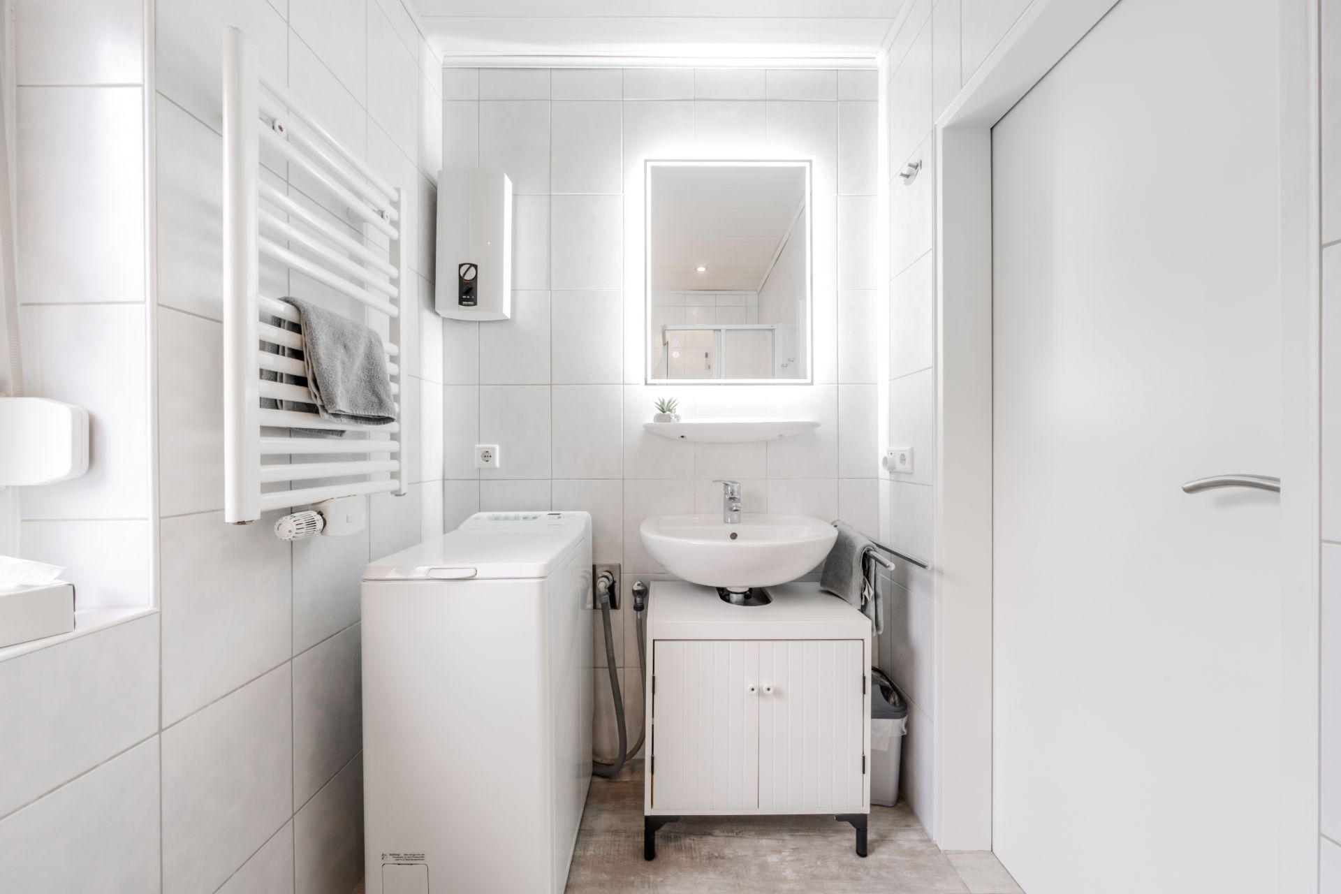 Modernes Badezimmer mit Waschmaschine, Waschbecken, beleuchtetem Spiegel und Handtuchheizkörper.