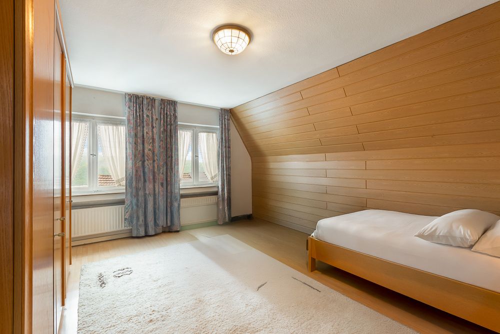 Schlafzimmer mit Einzelbett, Holzverkleidung an einer Wand, Teppich und drei Fenstern mit Vorhängen.
