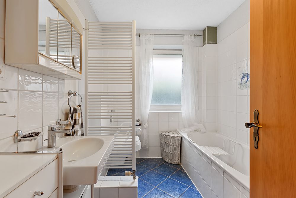 Helles Badezimmer mit Badewanne, Waschbecken, Fenster und blau gefliestem Boden.