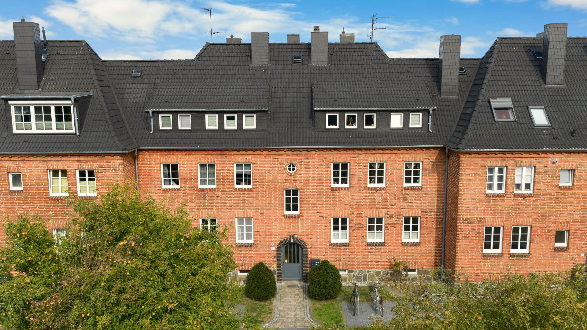 Backstein-Mehrfamilienhaus mit schwarzem Dach, mehreren Fenstern und grünem Vorgarten.
