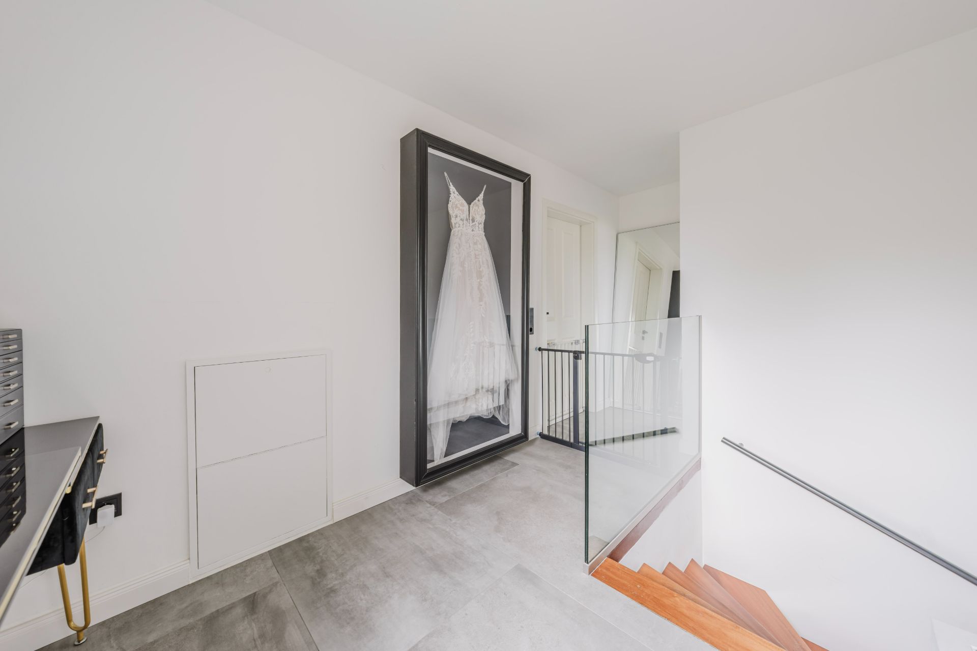 Heller Flur mit einem gerahmten weißen Kleid an der Wand, Treppe mit Glasgeländer.