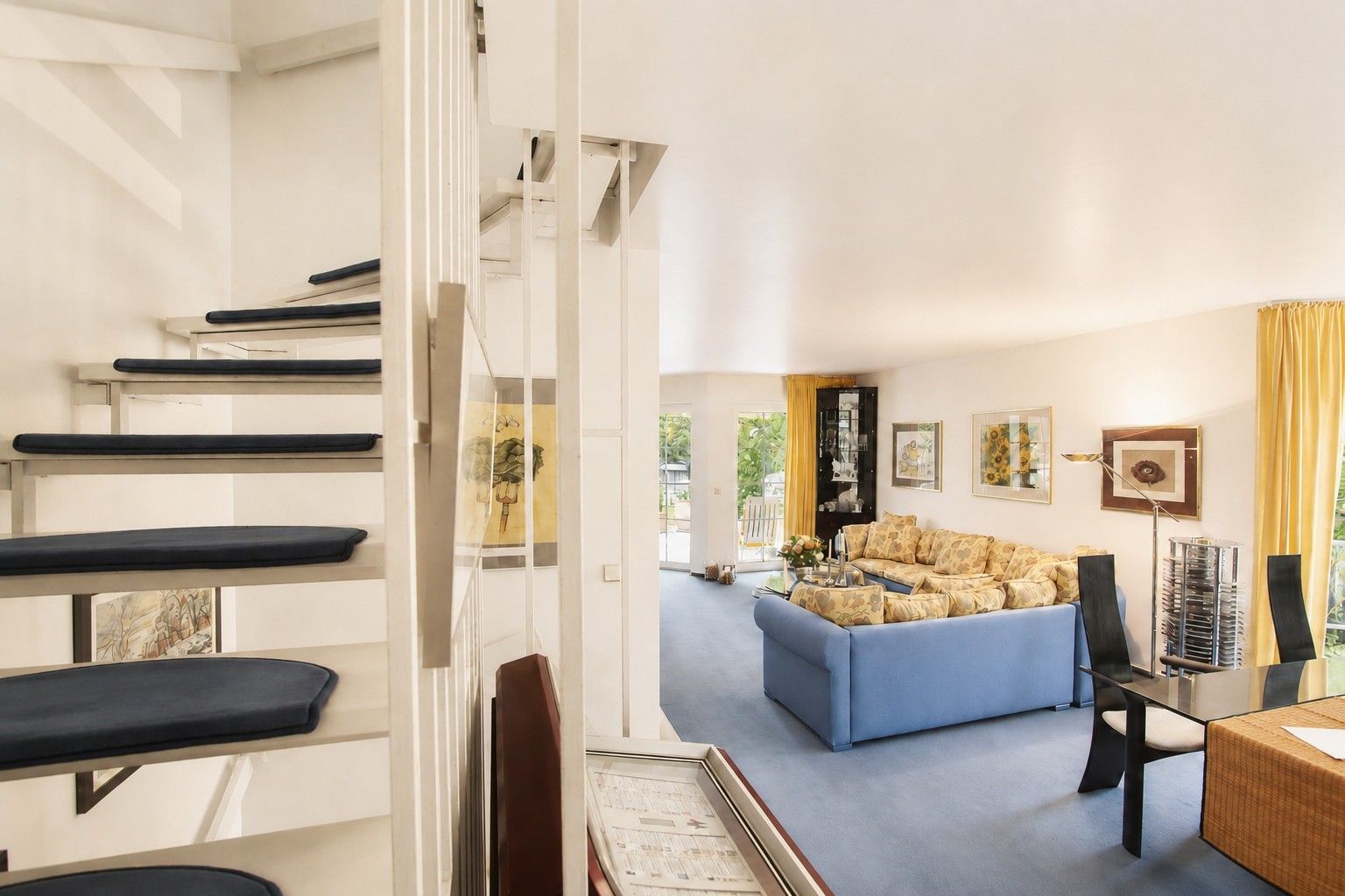 Modernes Wohnzimmer mit blauer Eckcouch, Glastisch und Treppe mit dunklen Stufen.