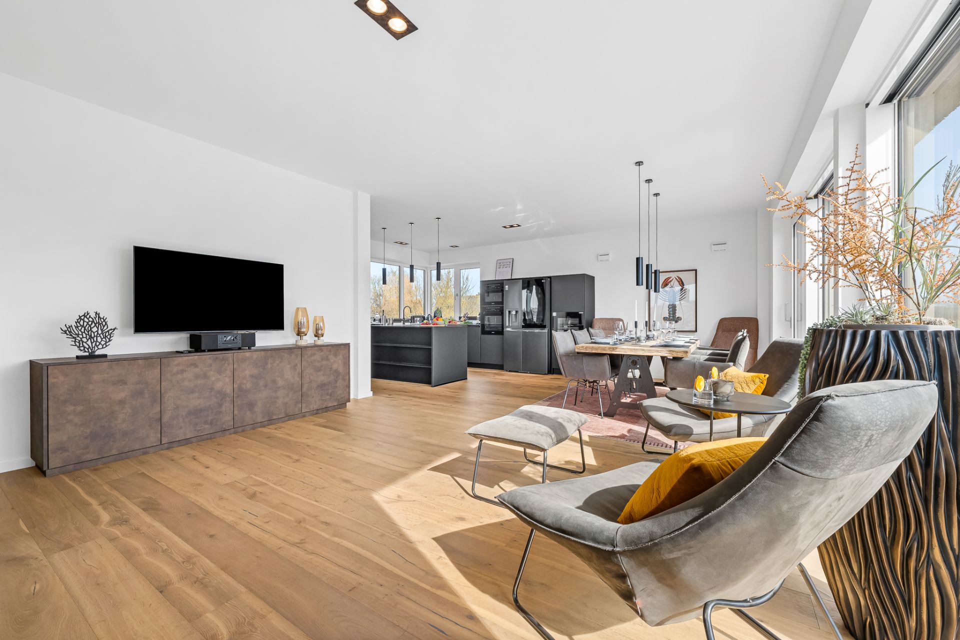 Helles Wohnzimmer mit Holzfußboden, Sesseln, Fernseher und Essbereich neben der offenen Küche.