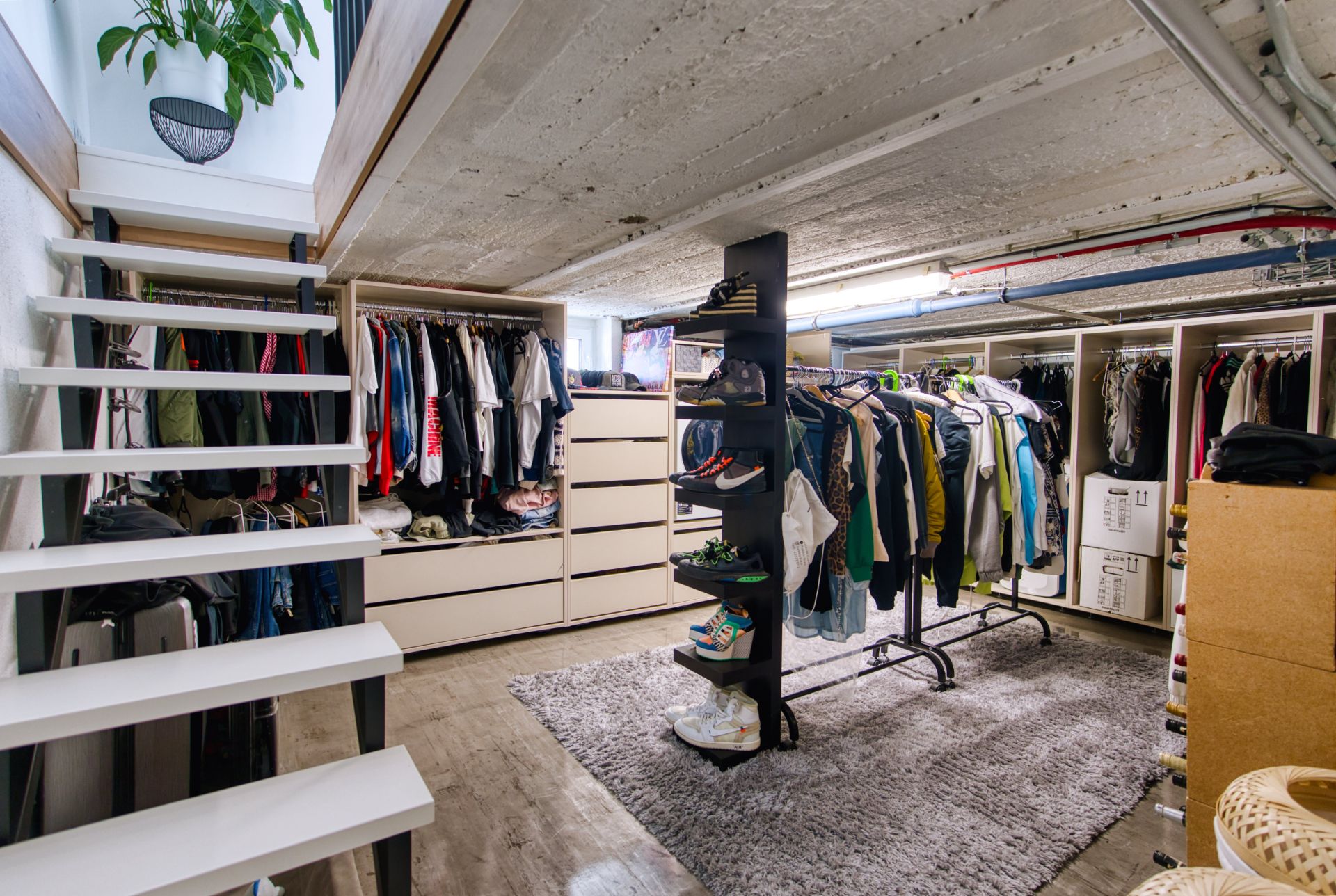 Begehbarer Kleiderschrank mit Regalen, hängender Kleidung, Kommoden und Teppich unter einer Treppe.