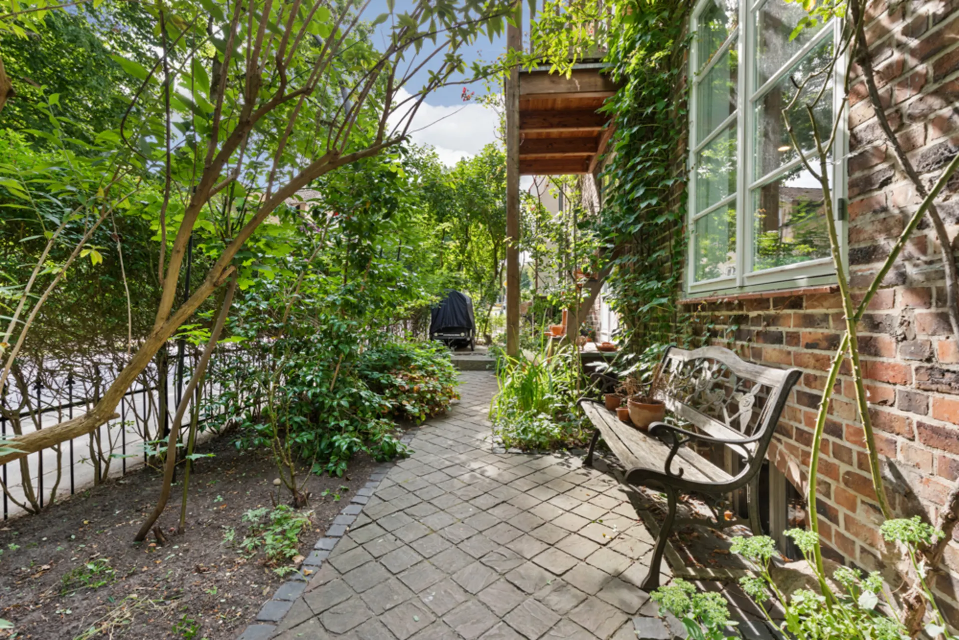 Gepflasterter Gartenweg neben Haus mit Bank, Pflanzen und gemauerter Hauswand.