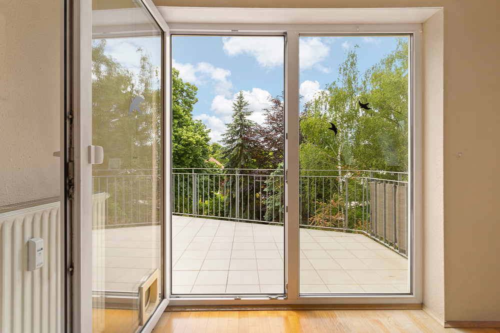 Offene Terrassentür mit Blick auf eine große geflieste Terrasse und grüne Bäume.