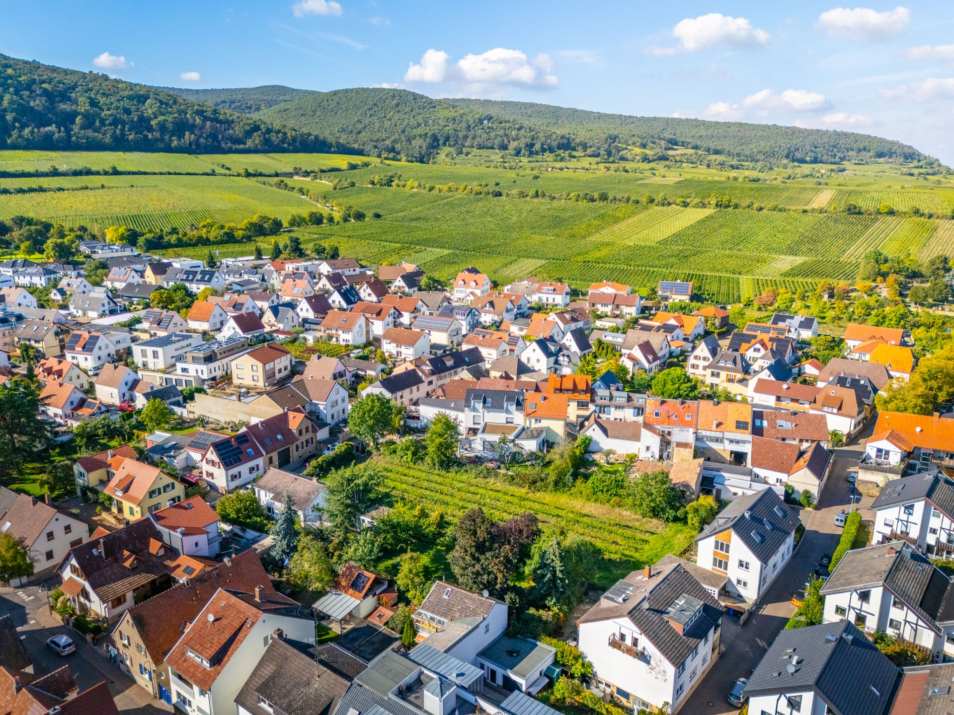 Luftaufnahme eines Wohnviertels mit roten Dächern und Feldern im Hintergrund.