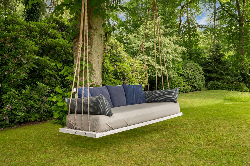 Große Hängeschaukel mit Kissen hängt an Seilen im Garten an einem Baum.
