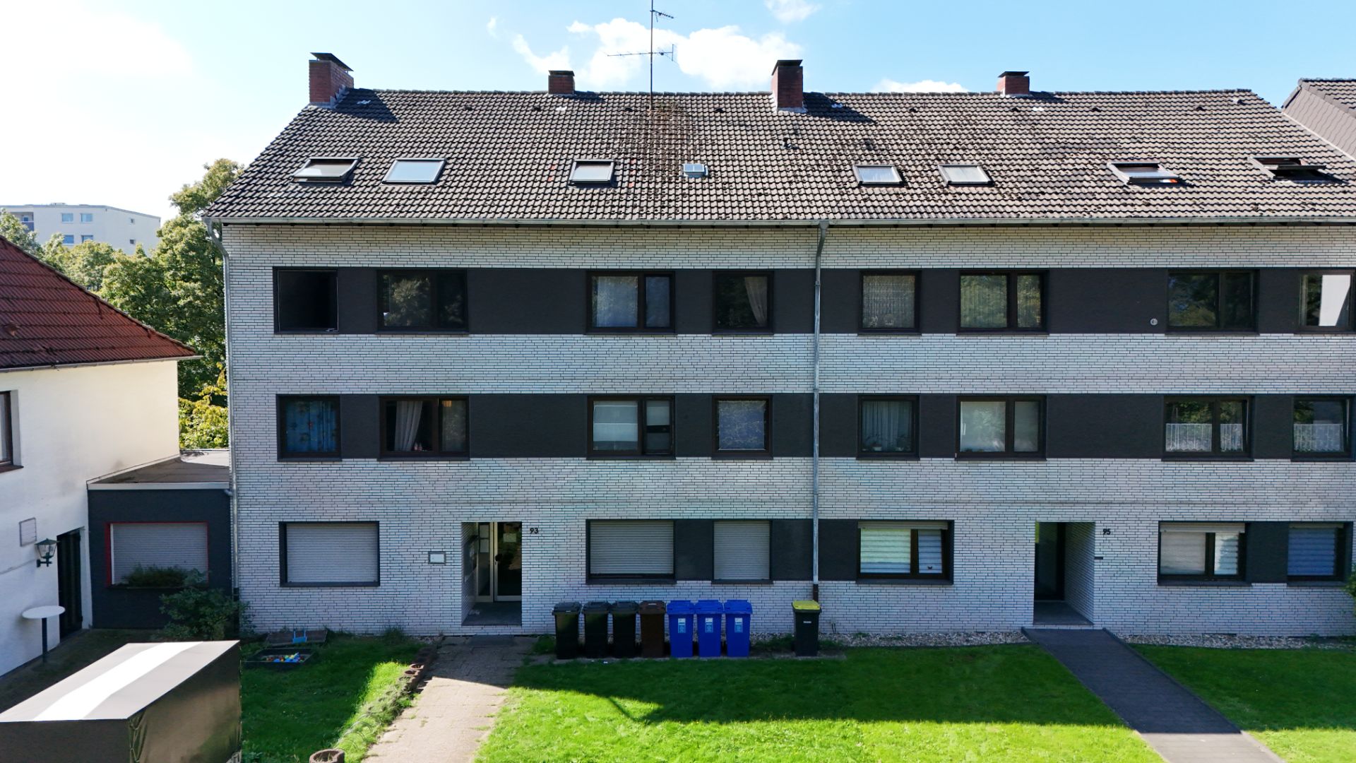 Dreistöckiges Wohnhaus mit mehreren Fenstern und Mülltonnen vor dem Eingang.
