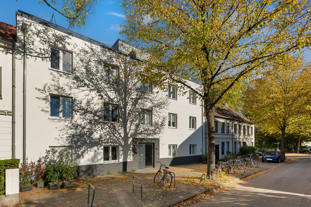 Modernes Wohnhaus mit weißen Wänden, Bäumen und parkenden Fahrrädern an einer ruhigen Straße.