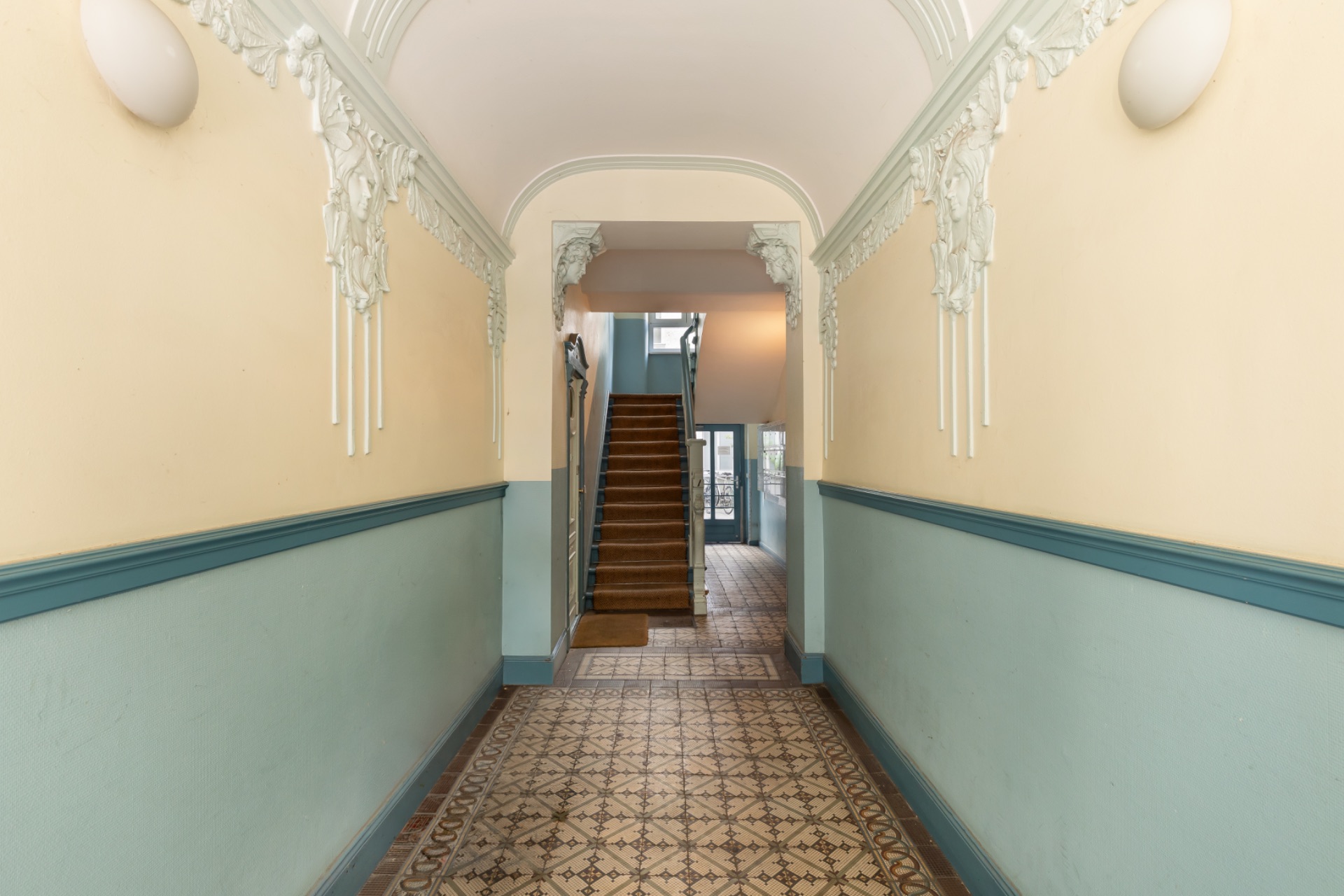 Heller Flur mit gemustertem Boden, Stuckverzierungen und einer Treppe am Ende.