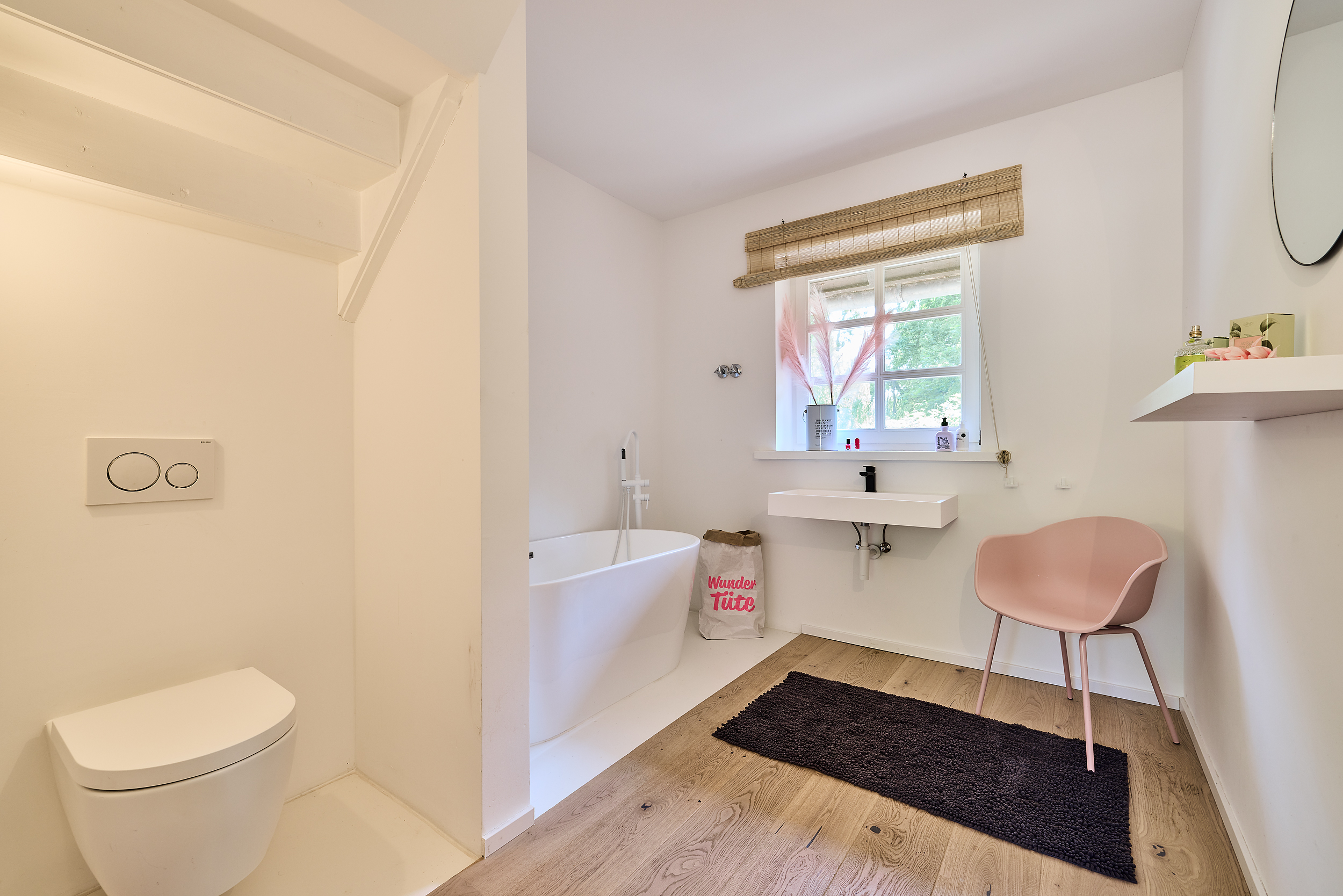 Helles Badezimmer mit Badewanne, WC, Waschbecken, rosa Stuhl und Fenster mit Blick ins Grüne.