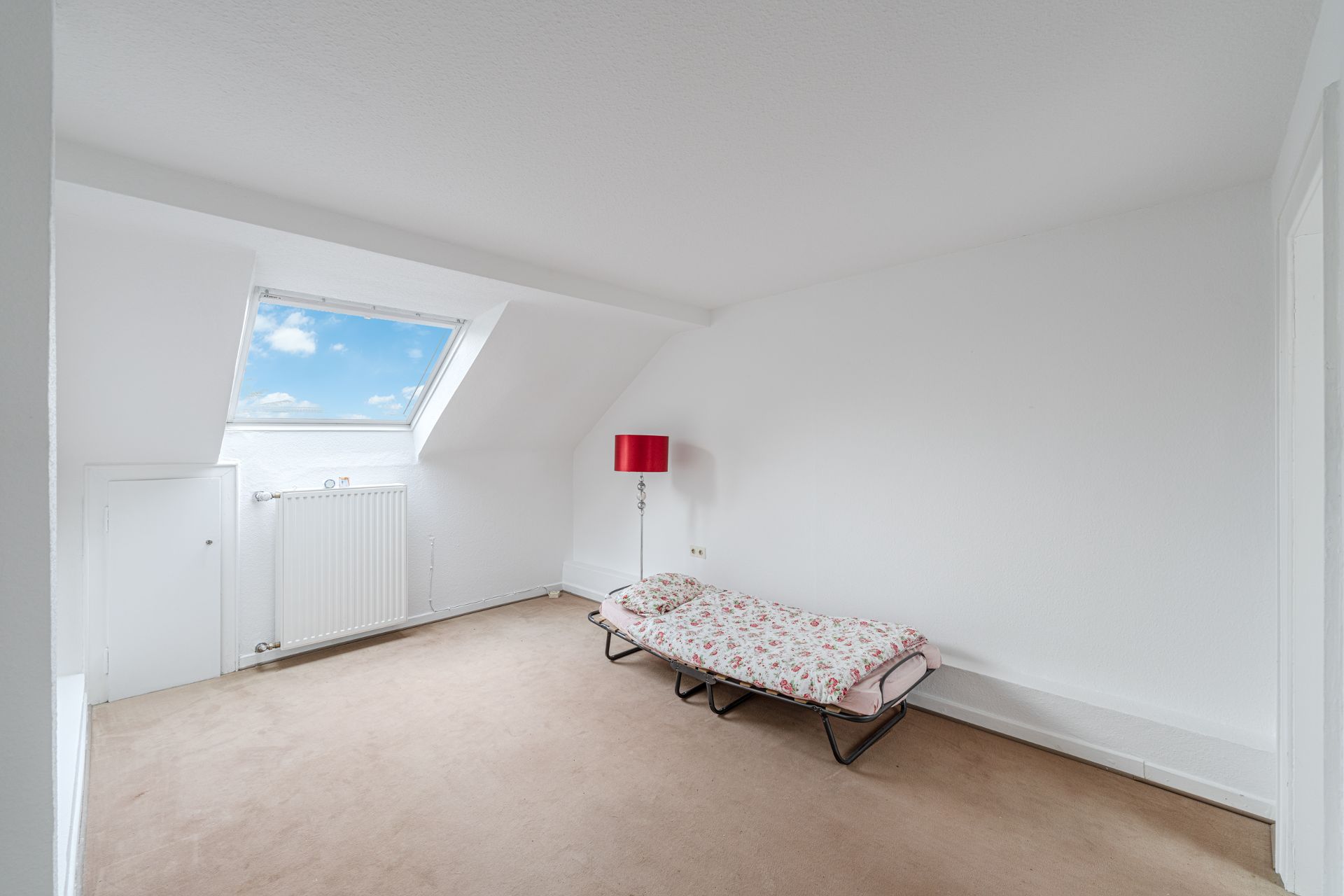 Helles Zimmer mit Dachfenster, Einzelbett und roter Stehlampe auf beigem Teppichboden.