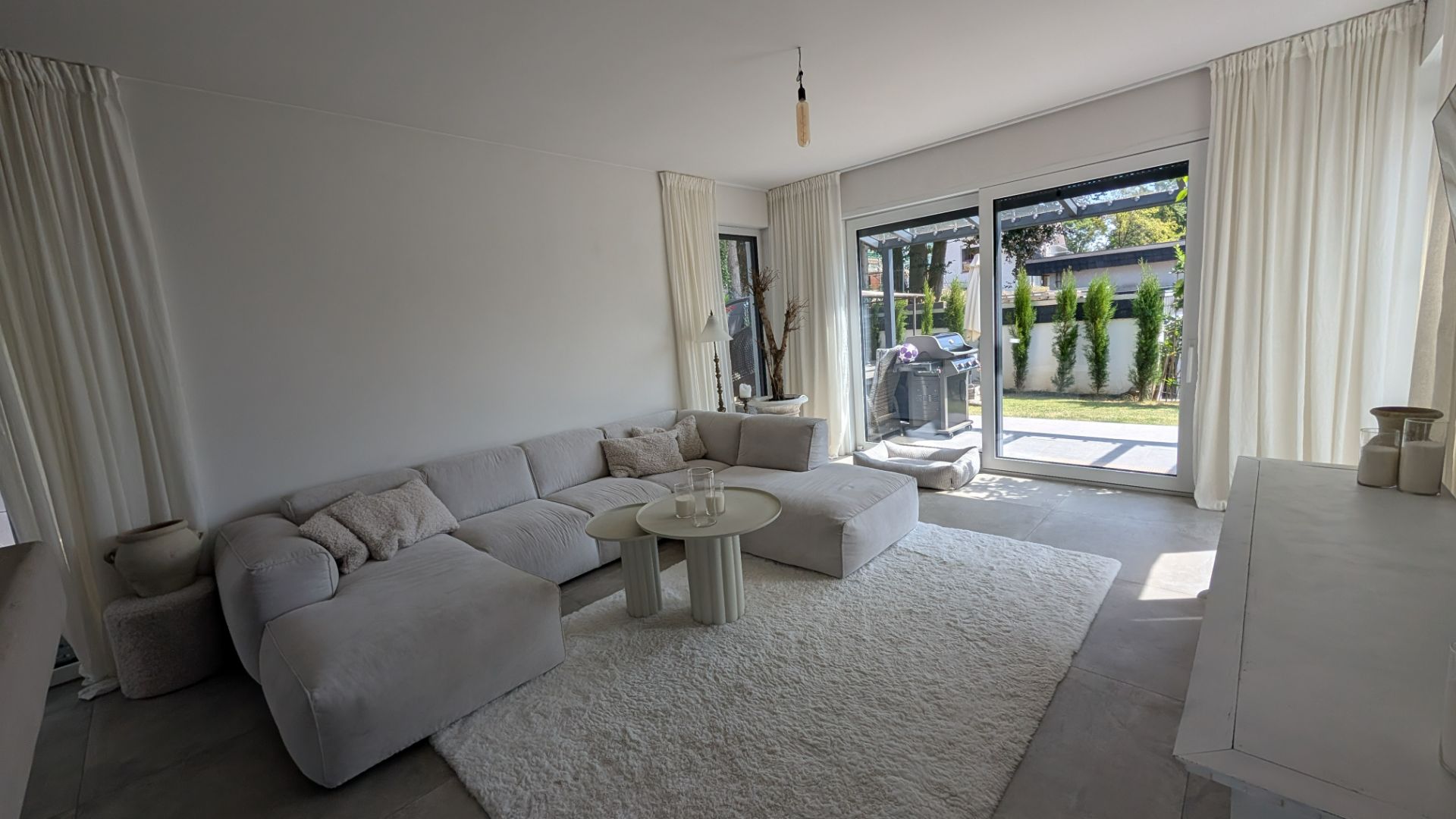 Helles Wohnzimmer mit Ecksofa, Teppich, Couchtisch und großen Fenstern mit Blick auf den Garten.