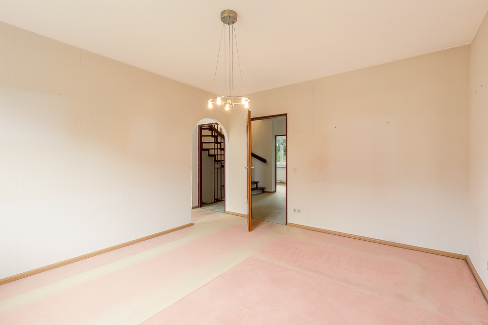 Leerstehendes Zimmer mit hellen Wänden, einem Kronleuchter und Blick auf einen Flur mit Treppe.